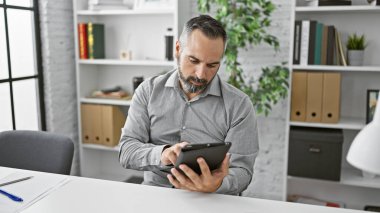 Sakallı, gri saçlı, modern ofis ortamındaki tablete odaklanmış yaşlı bir İspanyol..