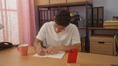 Kapalı ofis ortamında defterine yazı yazan genç İspanyol bir adam. Masasında bir telefon ve kahve fincanı var..