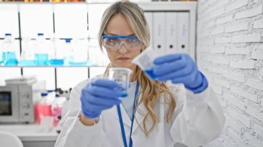 Laboratuvar önlüğü giymiş odaklanmış sarışın bir kadın modern bir laboratuvar ortamında deney yapıyor..