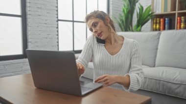 Konforlu bir oturma odasında akıllı telefonuyla sohbet ederken laptopuyla çalışan birden fazla işi olan genç bir kadın..