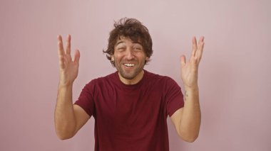 Sakallı, bordo tişörtlü, neşeli, genç, İspanyol bir adam pembe arka plan karşısında sevinçle heyecanını ifade ediyor..