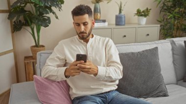 Yakışıklı İspanyol bir adam oturma odasında bir kanepede oturuyor, telefonuna dikkatle bakıyor, iç mekan bitkileri ve şık dekorlarla çevrili..