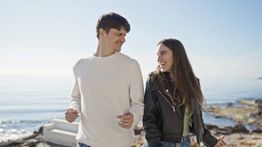 Gülümseyen bir çift, arka planda berrak bir okyanusla güneşli bir sahilde el ele tutuşuyor.