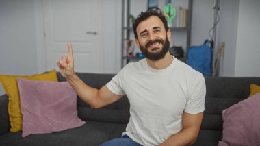 Gülümseyen sakallı bir adam renkli yastıklarla modern bir oturma odasını işaret ediyor..