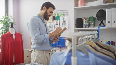 Sakallı bir adam terzide kumaş ve giysi raflarının arasında el işi ve tasarımı yansıtan notlar alır..
