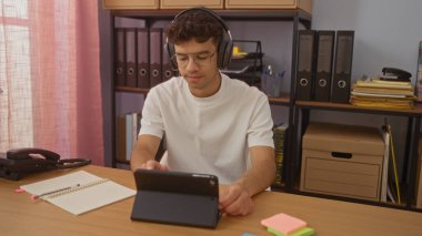 Genç, kulaklıklı bir İspanyol adam organize bir ofiste tablet üzerinde çalışıyor, ciltler ve kırtasiyelerle çevrili, odaklanmış ve üretken bir çalışma ortamı sunuyor..