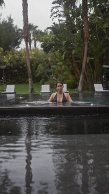 Düşünceli genç bir İspanyol kadın tropikal bale bitkileriyle çevrili açık havuzda sakin bir anın tadını çıkarıyor..