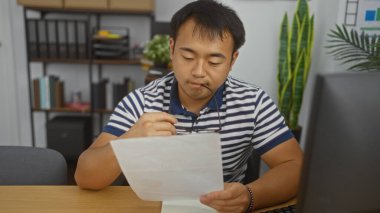 Ofiste çalışan yakışıklı genç Çinli adam, elinde gözlüklerini tutarken dikkatle bir belge okuyor. Asyalı yetişkin erkek, organize kitap rafları ve bitkilerle kapalı bir iş yerinde odaklanıyor..