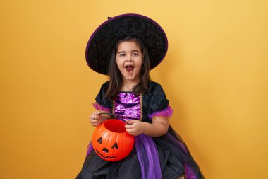 Cadı kostümü giymiş tapılası İspanyol kız, ağzı açık inanamıyor, korkmuş ama heyecanlı balkabağı sepeti tutuyor, sarı arka planda izole edilmiş.