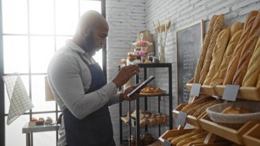 Sakallı ve kel kafalı genç bir adam arka planda ekmek ve hamur işleri rafları olan bir pastanede tablet kullanıyor.