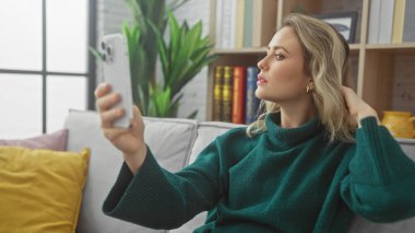 Güzel sarışın bir kadın, modern bir yaşam tarzını yansıtan rahat bir oturma odasında selfie çekiyor..