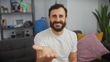 Beyaz tişörtlü, gülümseyen sakallı adam sıcak bir oturma odasında el sıkışmak için elini uzatıyor..
