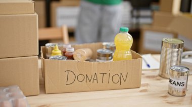 Blurred young man volunteering in a warehouse, facing a donation box with food and beverages ready for charity. clipart