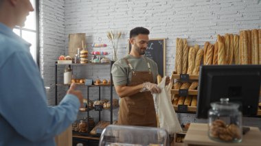 Bir pastanede iki adam önlük ve eldiven takmış ekmek paketlerken diğer adam tezgahın öbür ucundan birini işaret ediyor.