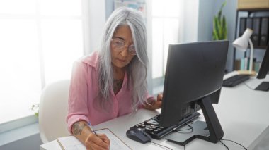 Ofis masasında notlar yazan, parlak ve modern ofis ortamında bilgisayar üzerinde çalışırken odaklanan ve gözlük takan olgun bir kadın..