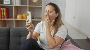 Yorgun bir kadın akıllı bir telefon tutarken esniyor, rahat bir oturma odasında gri bir kanepede oturuyor..