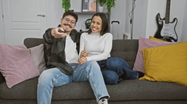 Oturma odasındaki kanepede yan yana uzanmış gülümseyen bir çift, kamerayı işaret ederek, aşkı ve arkadaşlığı somutlaştırıyor..