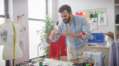 Terzide sakallı bir adam, arka planda tasarımları olan bir elbise formundaki resmi çekmek için akıllı telefon kullanıyor..