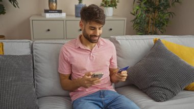 Modern oturma odasındaki koltukta otururken elinde telefon ve kredi kartı olan genç adam online alışveriş ya da bankacılık...