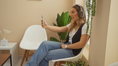 Kulaklık takan güzel sarışın kadın kapalı bir bekleme odasında oturup telefonuyla selfie çekiyor..