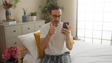 Orta yaşlı İspanyol bir adam, rahat bir yatak odasında, çiçekleri ve kitapları olan bir yatakta otururken akıllı telefonuyla video görüşmesi yapıyor..