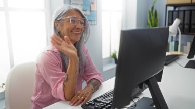 Ofis ortamında video konferans odasında konuşan bir kadın kameraya güvenle gülümseyen, gözlüklü ve pembe tişörtlü, gri saçlı, modern ofis ekipmanlarıyla çevrili..