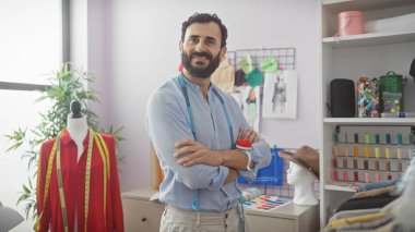 Modeller, kumaşlar ve ipliklerle çevrili bir tasarım odasında kolları çapraz duran kendine güvenen erkek terzi..