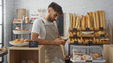 Fırında sakallı genç bir adam, evde dikiliyor ve telefonunu kontrol ediyor. Önlük takıyor, çeşitli fırın malzemeleri sergiliyor..