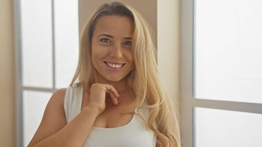 Sarışın kadın sıcak bir şekilde gülümsüyor. Parlak, modern bir iç mekan ortamında. Günlük zarafet ve yakınlık yayıyor..