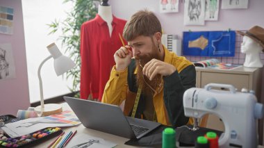Sakallı, odaklanmış kızıl saçlı bir adam renkli bir terzide dikiş ekipmanları arasında dizüstü bilgisayar kullanarak çalışıyor..