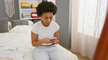 Afro-Amerikalı bir kadın, yatak odasında, kapalı bir yatakta oturmuş, akıllı telefonuyla kıvrılıyor.