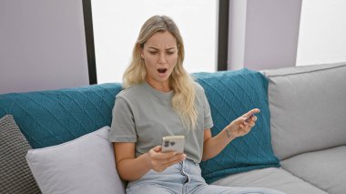Şaşırmış genç bir kadın evdeki koltukta akıllı telefon kullanıyor, modern, gelişigüzel, teknoloji, yaşam tarzı, kadın, kapalı alan, beyaz, ifade, iletişim..