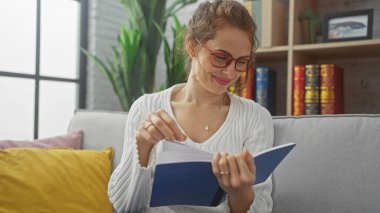 Genç bir kadın güzel dekore edilmiş oturma odasında rahat bir kanepede kitapçık okuyor..