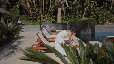 Beyaz elbiseli genç bir kadın yemyeşil yaprakların arasında güneşlenmek için havuz hazırlıyor..