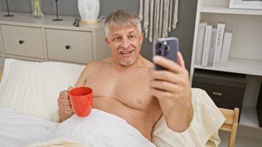 Orta yaşlı bir adam yatak odasında kahveyle selfie çeker. Evde rahat bir yaşam tarzı sergiler..