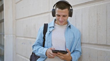 Genç bir İspanyol adam, güneşli bir şehir sokağında kulaklıklı bir tablet kullanırken dışarıda durup gülümsüyor..