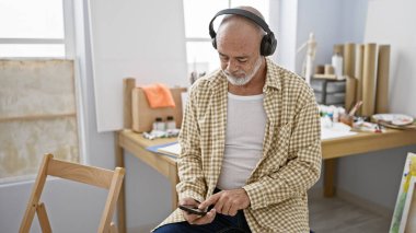 Sakallı ve kulaklıklı olgun bir adam stüdyoda içinde sanat malzemeleri olan akıllı bir telefon kullanıyor..