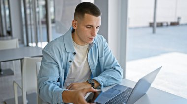 Odaklanmış genç bir adam, modern bir üniversite kütüphanesi ortamında dizüstü bilgisayar üzerinde çalışıyor, günlük tarz ve akademik çalışkanlığın bir karışımını somutlaştırıyor..