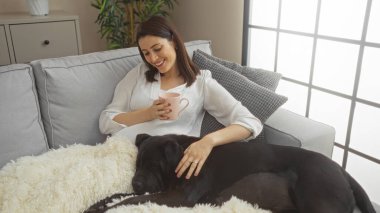 Genç bir İspanyol kadın labrador köpeği ve yumuşak ışıklandırmalı, rahat, modern bir oturma odasında bir fincan kahveyle koltukta rahatlar..