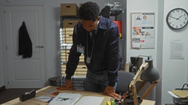 Odaklanmış, rozetli bir Afrikalı Amerikalı polis karakolunun soruşturma odasında belgeleri inceliyor..