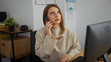 Sarışın, genç, ofiste bir kadın, bilgisayarının başında çalışırken konuşuyor, etrafı ofis malzemeleri ve dekorla çevrili..