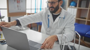 Sakallı, orta yaşlı bir erkek doktor hastane odasında dizüstü bilgisayarla çalışıyor.