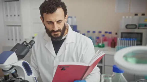 Stock image A middle-aged, bearded man in a lab coat reads a book in a well-equipped laboratory, indicating research or education.