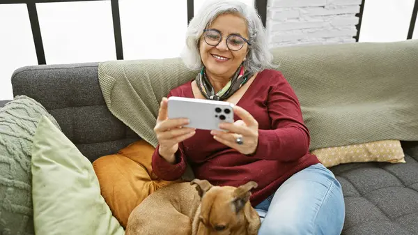 Gülümseyen gri saçlı bir kadın, evinde yanında kahverengi bir köpekle birlikte renkli yastıklı bir akıllı telefon kullanıyor..
