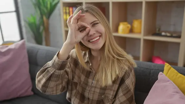 stock image Smiling young woman making okay sign over eye, relaxed at home on cozy sofa, playful mood in a comfortable interior.