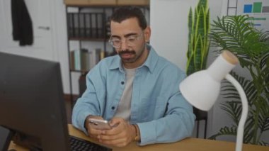 Ofisteki masada bilgisayar ve bitkilerle akıllı telefon kullanan genç bir adam modern ve profesyonel bir çalışma ortamı yaratıyor..