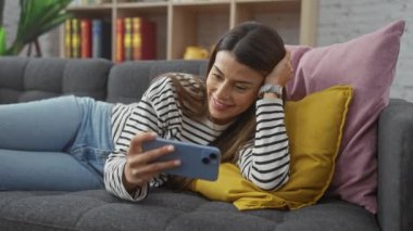 Gülümseyen İspanyol bir kadın, akıllı telefonuyla meşgul bir şekilde evdeki kanepeye uzanır.