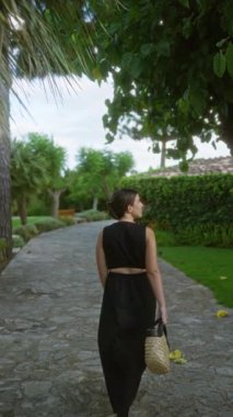 İspanya, Mallorca 'da taş bir yolda yürüyen, siyah bir elbise giyen ve yemyeşil bir yerde ayakkabılarını taşıyan bir kadın.