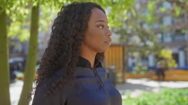 Güzel, Afro-Amerikan bir kadın şehir parkında duruyor, kıvırcık saçlı ve siyah bluzlu..
