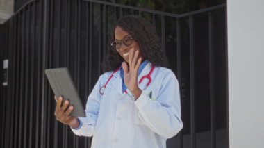 Kıvırcık saçlı ve gözlüklü gülümseyen Afro-Amerikalı bir kadın doktor, şehir merkezindeki bir hastanenin yanında beklerken tablet kullanarak bir video görüşmesi yapıyor..
